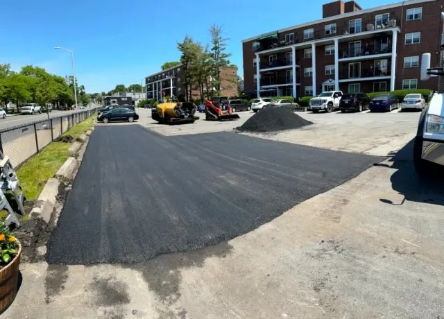 Freshly paved asphalt in a parking lot, construction equipment in the background. Buildings and cars are visible.