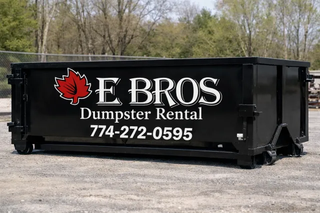 Black dumpster with logo and text for E BROS Dumpster Rental, including a red leaf graphic and contact number, on a gravel surface with trees in the background.