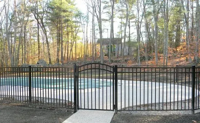 Black metal fence surrounds a swimming pool with a gate, trees in the background.