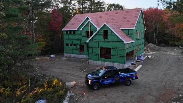A dark pickup truck parked in front of a new two-story house under construction, surrounded by autumn trees.