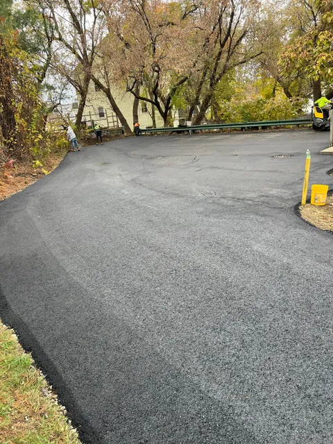Newly paved asphalt driveway curving toward trees and a house on a fall day.