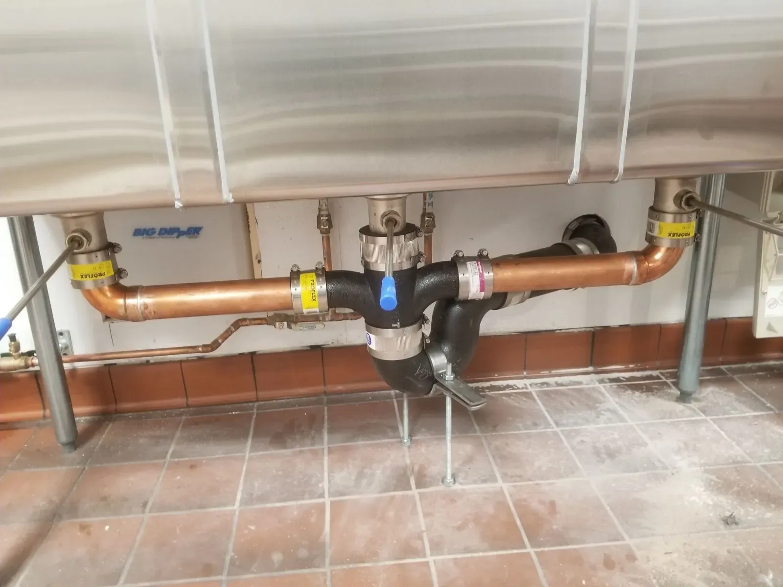 A stainless steel sink drain setup with copper pipes, labeled valves, and a central trap is expertly installed by a plumber in Plymouth and Suffolk County, MA. It’s mounted against a tiled wall above a brown tile floor.
