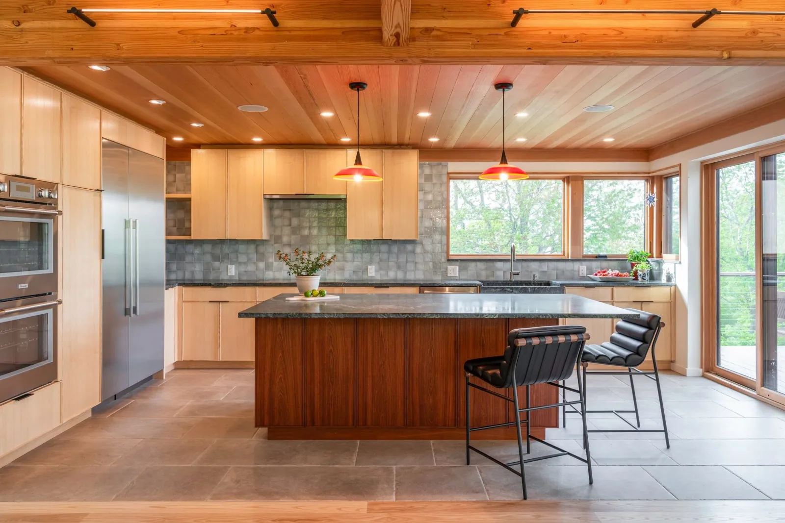 Modern kitchen with wooden ceiling and cabinets, gray tiled backsplash, large windows, black countertop, kitchen island with wooden base, two black bar stools, and potted plants.