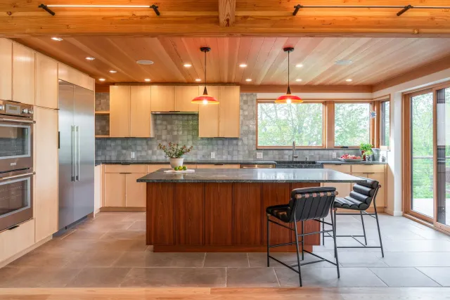 Modern kitchen with wooden ceiling and cabinets, gray tiled backsplash, large windows, black countertop, kitchen island with wooden base, two black bar stools, and potted plants.