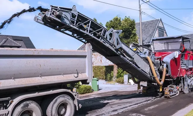 An asphalt milling machine transferring removed pavement material onto a dump truck via conveyor belt during a road resurfacing project on a residential street.
