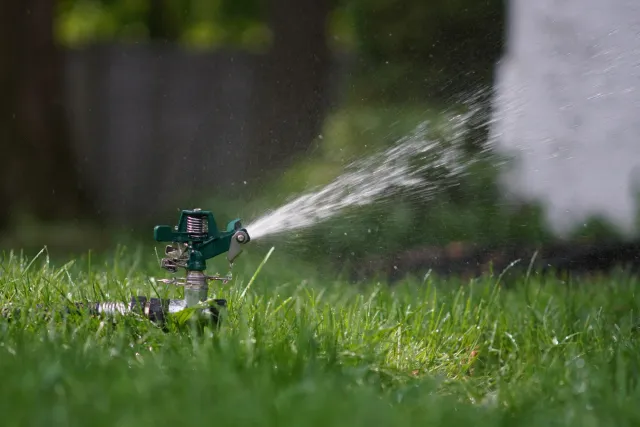 A green lawn sprinkler watering grass, with water spraying out in a fine mist and droplets visible in the air.