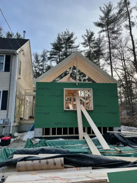 A wooden home addition under construction with green sheathing, a gable roof frame, and temporary support beams.