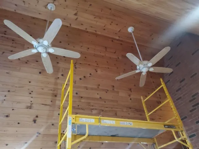 Two ceiling fans installed on a wood plank ceiling; a yellow scaffold stands beneath the fans.