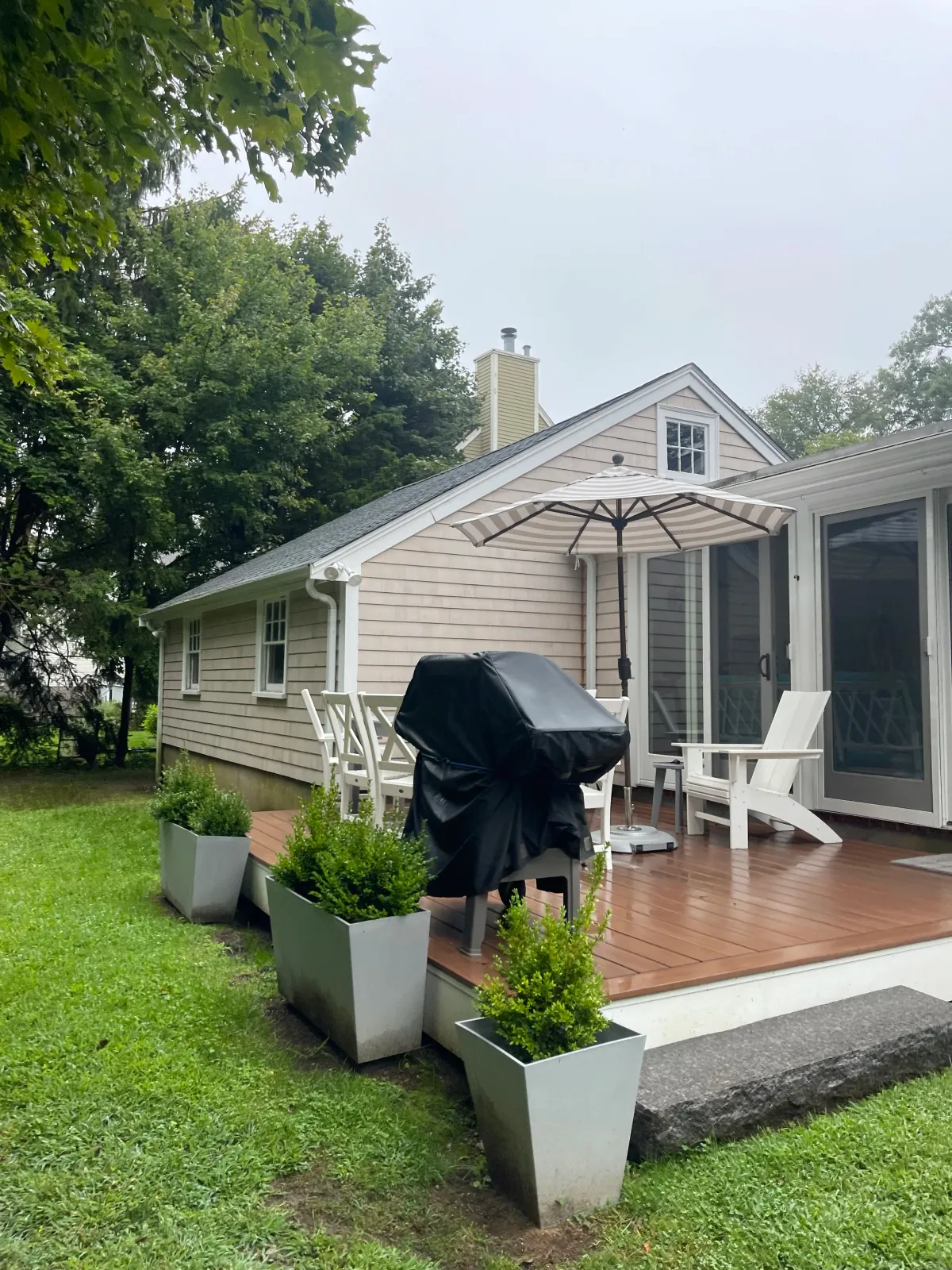 Backyard patio with outdoor furniture, a covered grill, potted plants, a deck, and a house with large windows and a sliding door, surrounded by green grass and trees.