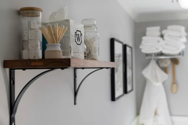 A bathroom wall with a wooden shelf holding glass jars and toiletries, next to framed art and stacked towels on a rack.