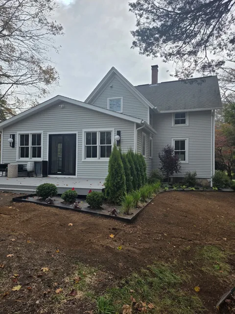 A light gray house with a porch and garden bed in front of an overcast sky.