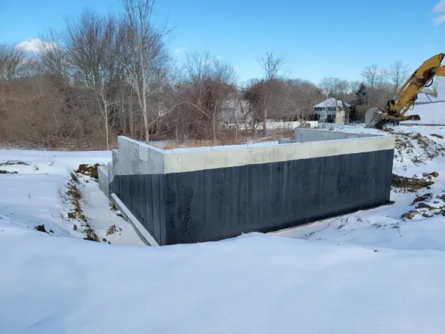 Exterior of concrete foundation coated with black material, surrounded by snow, in a construction site.