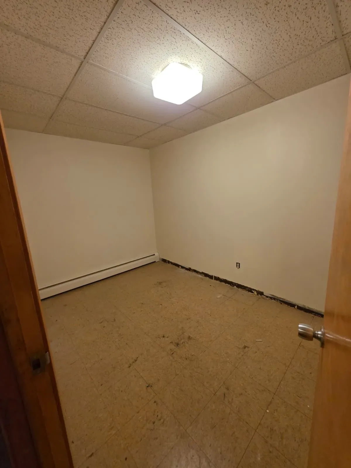 Empty beige room with a tiled ceiling, hanging light, and a bare floor viewed through an open door