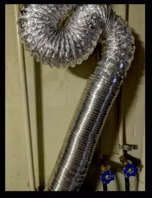 A flexible, metallic dryer vent hose loops down against a wall above two blue-handled water valves.