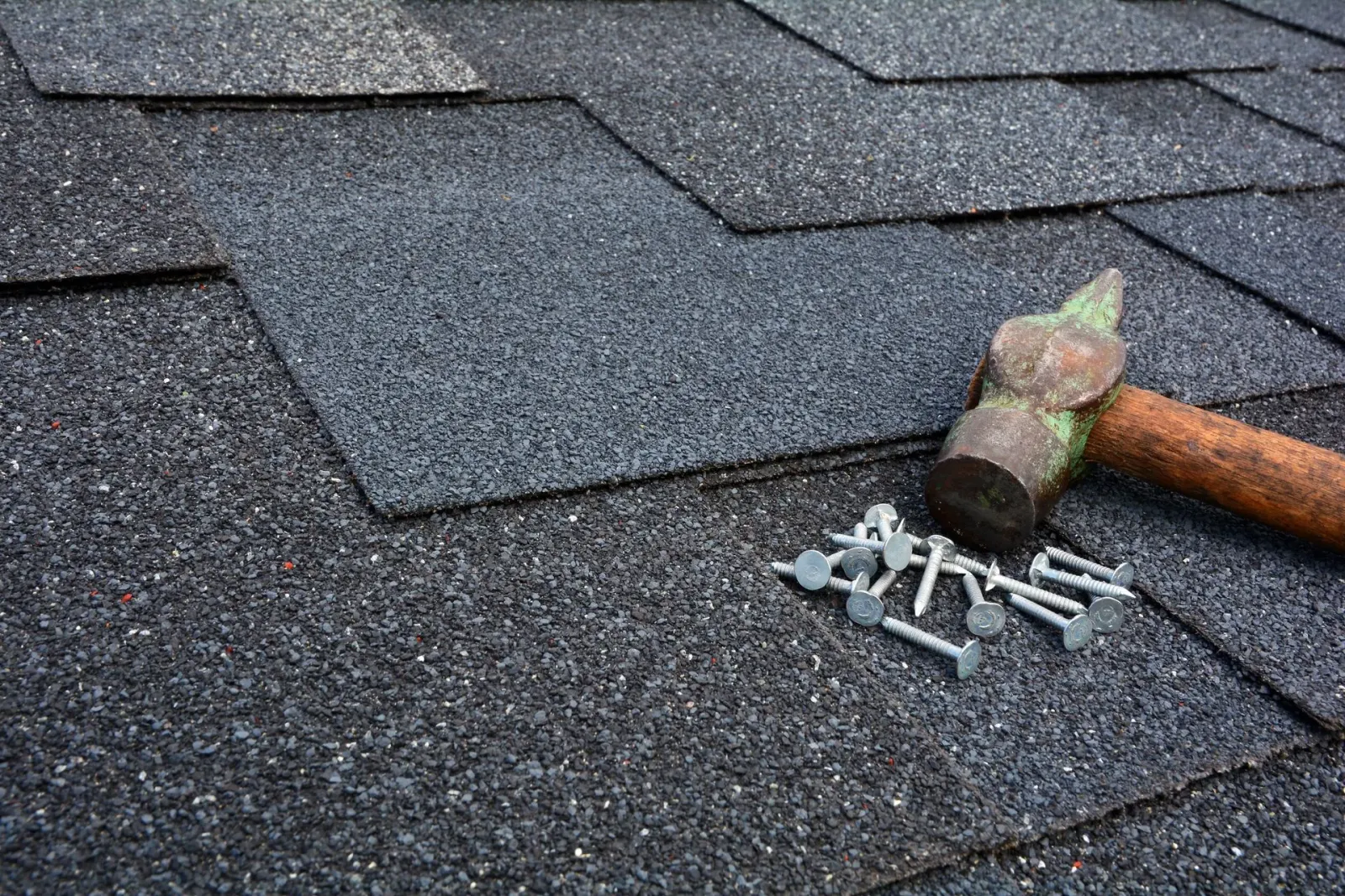 A hammer and a small pile of roofing nails resting on a dark gray asphalt shingle roof.