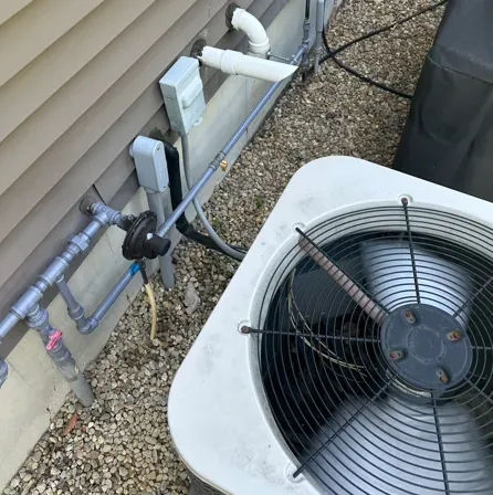 Outdoor HVAC unit beside house siding, with exposed pipes and electrical boxes on a gravel bed.