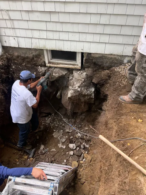 Man using a jackhammer to break up rocks near a house foundation
