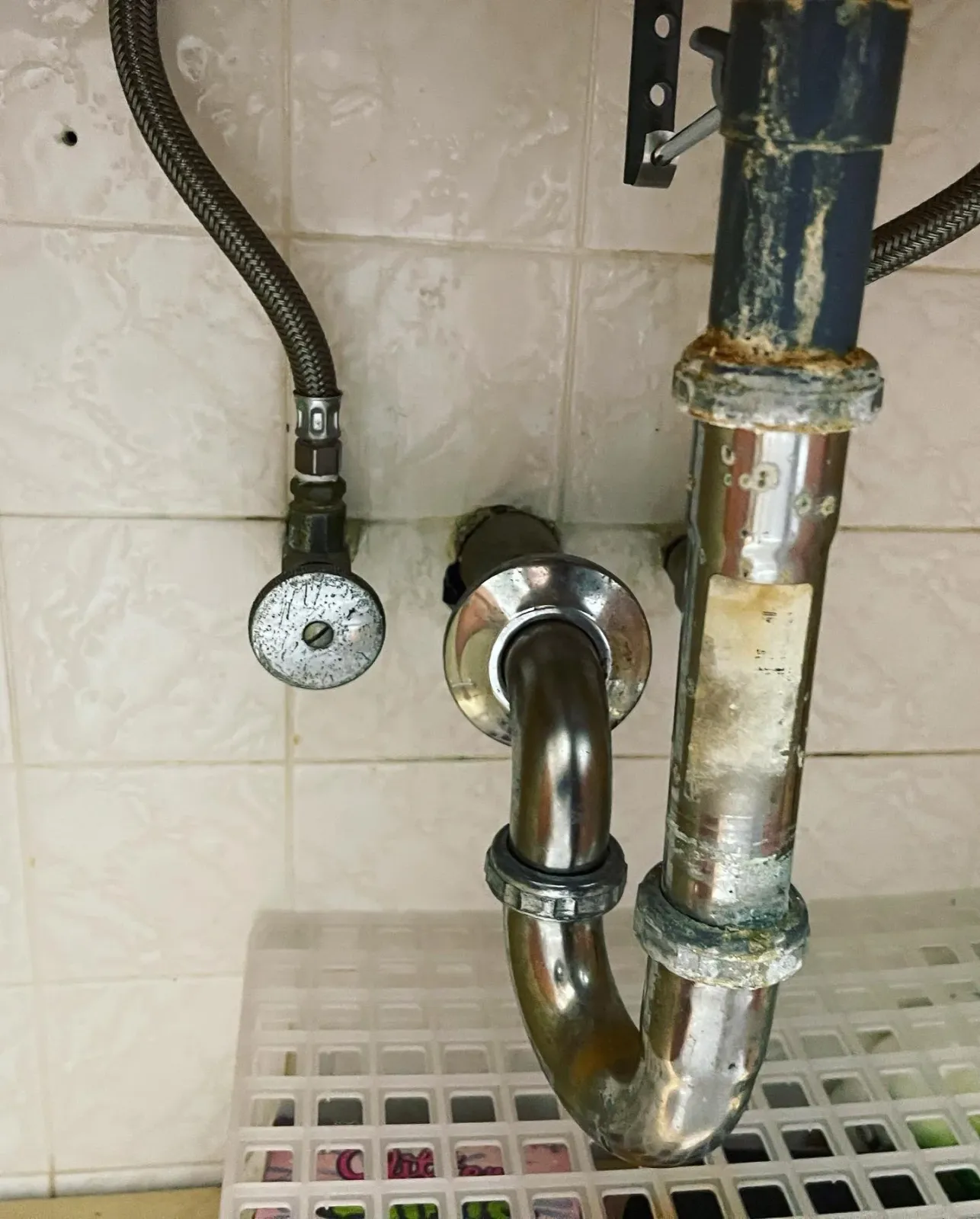 Underneath a kitchen sink, there is a metal pipe connected to the drain and water supply lines. The pipes are rusty and stained, with a flexible black hose nearby, all mounted on a beige tiled wall.