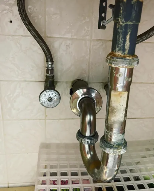 Underneath a kitchen sink, there is a metal pipe connected to the drain and water supply lines. The pipes are rusty and stained, with a flexible black hose nearby, all mounted on a beige tiled wall.