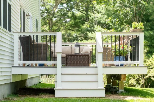 A white backyard deck with outdoor furniture, including a wicker sofa, a wicker chair, and a lantern on a coffee table, surrounded by green trees and plants.