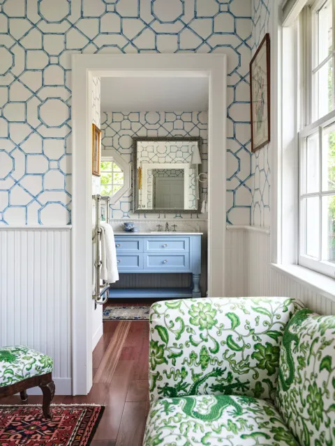 View of a bathroom seen through a doorway from a room with green and white patterned furniture, featuring a blue vanity, framed mirrors, and patterned wallpaper.