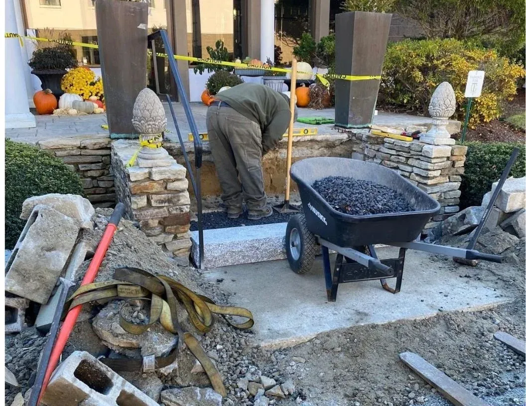 A person working on constructing or repairing a stone or brick entranceway, with tools and construction materials around, and pumpkin and autumn decor in the background.