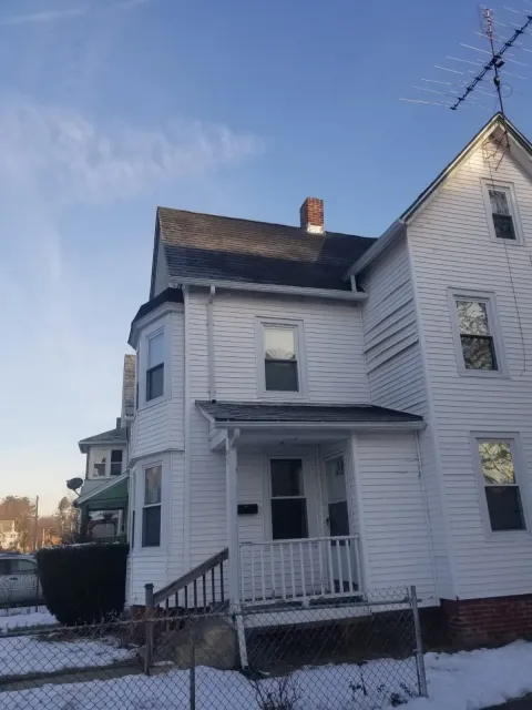 White, multi-story house with a front porch, fenced yard, and snow on the ground under a clear blue sky.