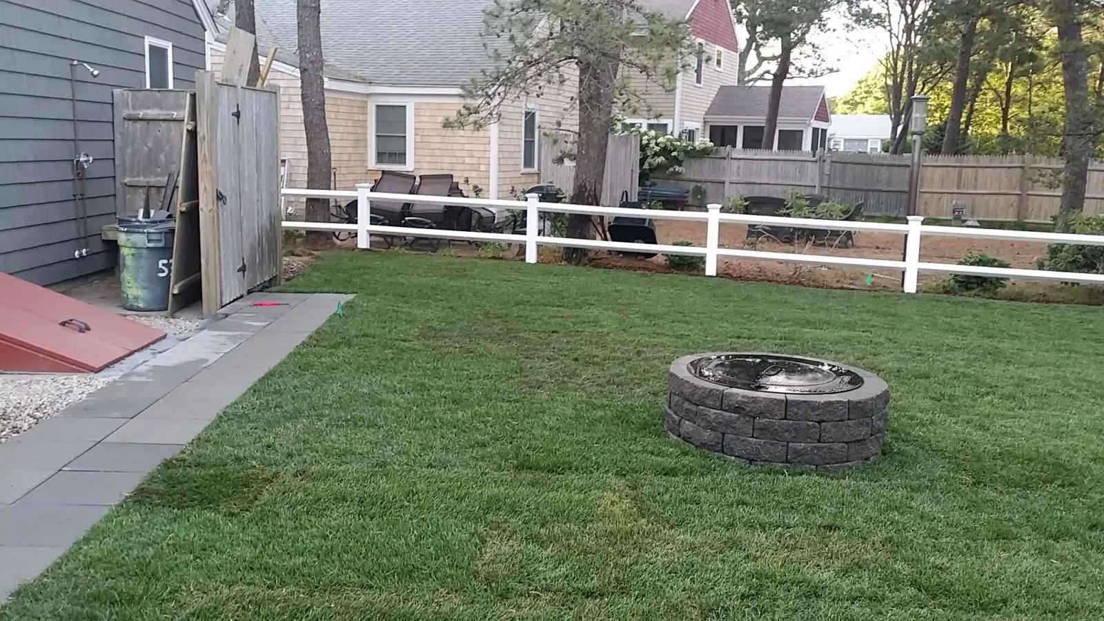 Backyard with green lawn, fire pit, white fence, and outdoor seating area beside a house with gray siding.