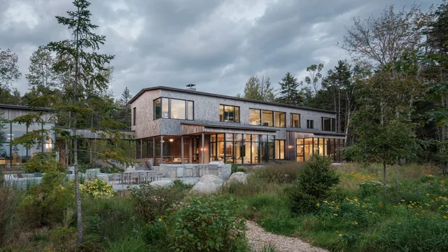 Modern wooden house with expansive windows nestled in lush Maine forest.