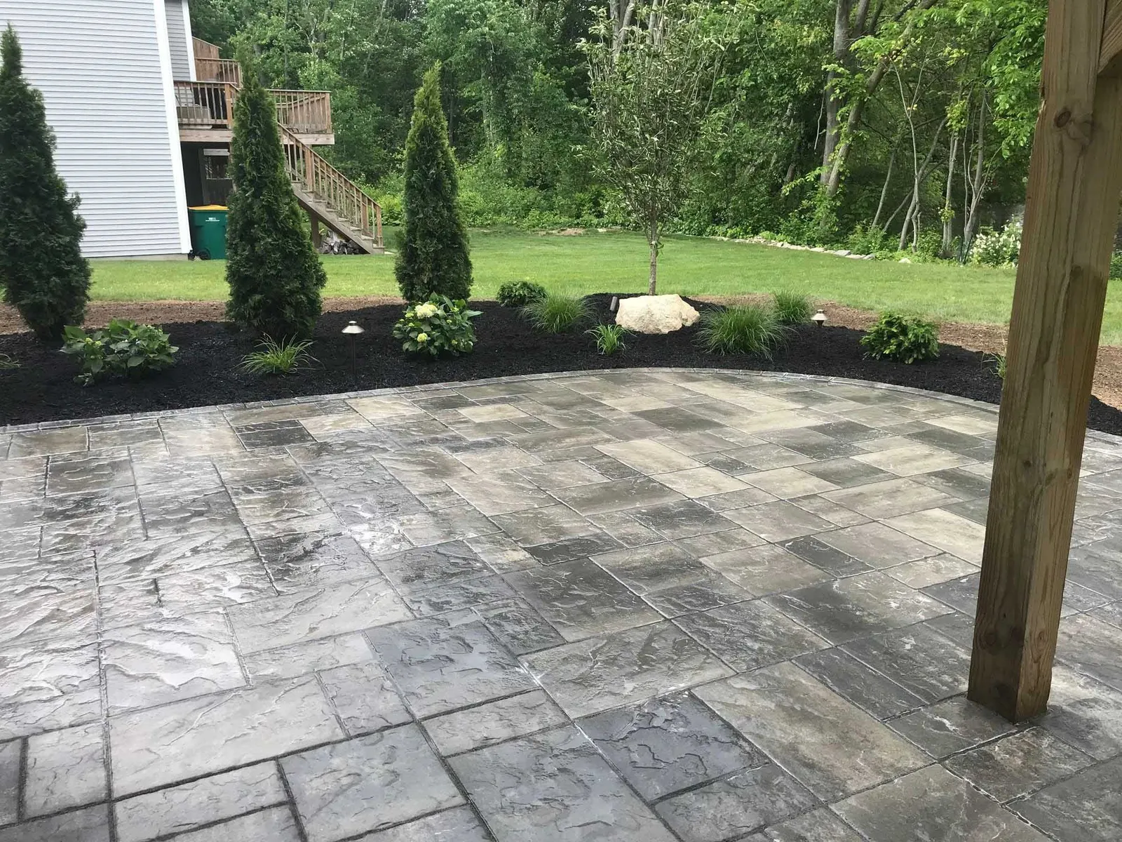 Paver patio with landscaping, featuring dark mulch, bushes, and trees. Wooden post on right.