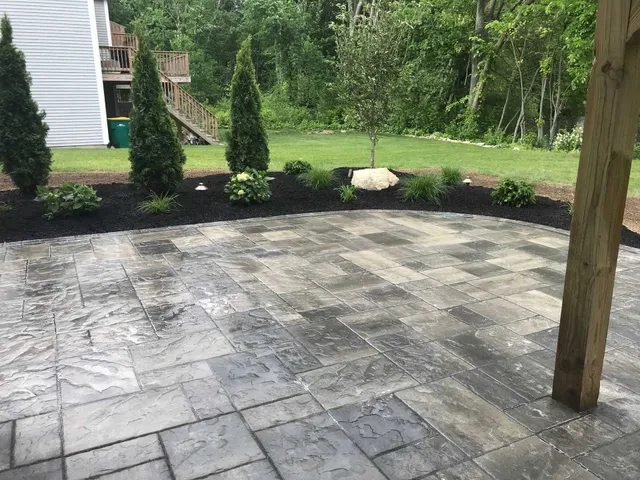 Paver patio with landscaping, featuring dark mulch, bushes, and trees. Wooden post on right.