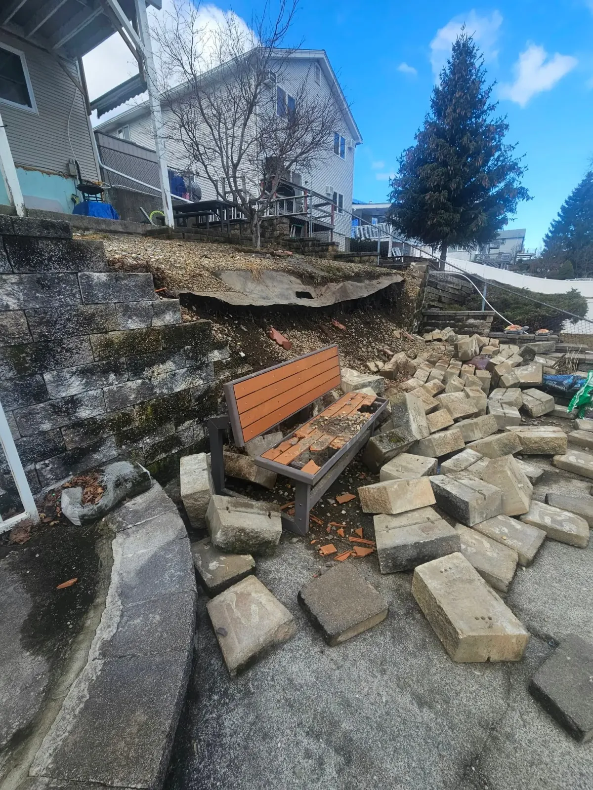 Collapsed retaining wall with scattered concrete blocks and a bench in a residential yard.