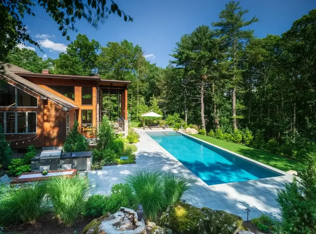 Modern wooden house with glass walls framing a long rectangular pool and patio seating amid a forest backdrop.