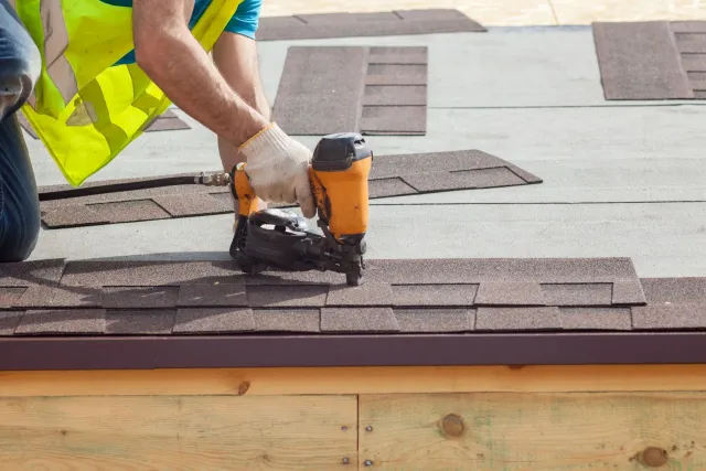 A worker in a yellow safety vest uses a nail gun to attach brown asphalt shingles to a roof.