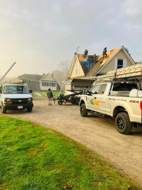 Workers and trucks at a storm-damaged house with a torn roof and debris in the driveway