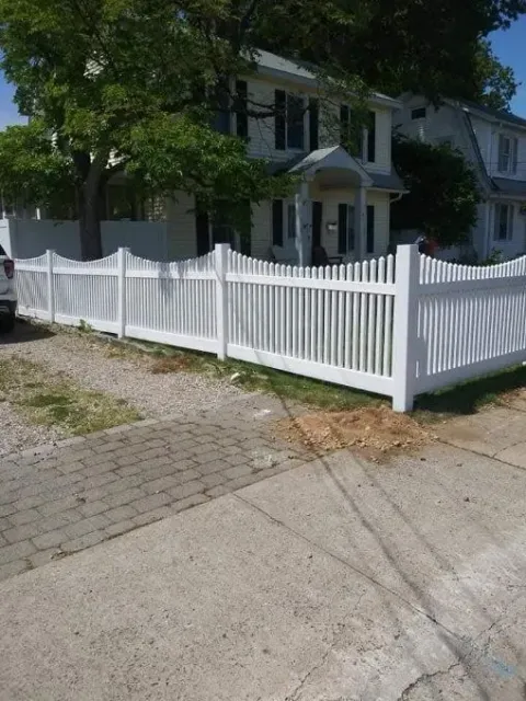 White picket fence in front of a two-story house, gravel driveway, and brick sidewalk.