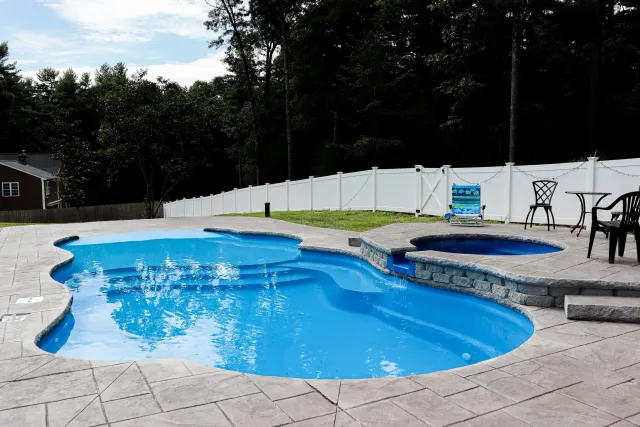 Backyard swimming pool with a hot tub, surrounded by pavers, with chairs and a fence in the background.