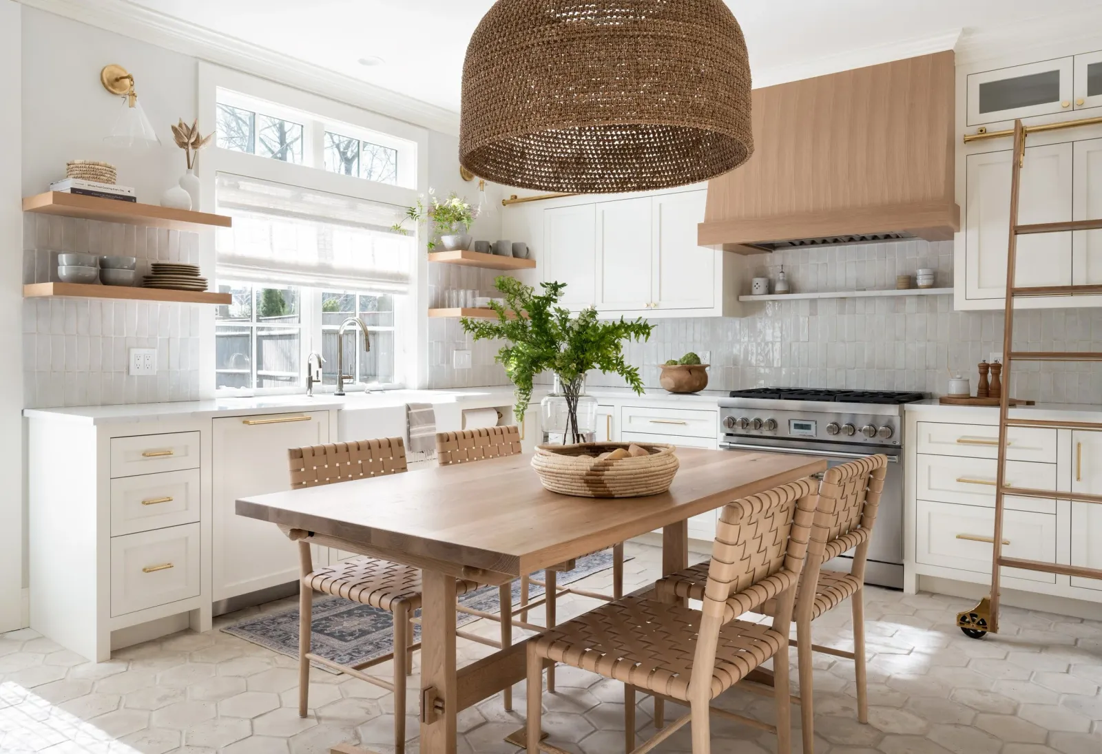 Bright kitchen with white cabinetry, wooden accents, and a wooden dining table with woven chairs. Large window, green plants, and open shelves with dishes and decor.