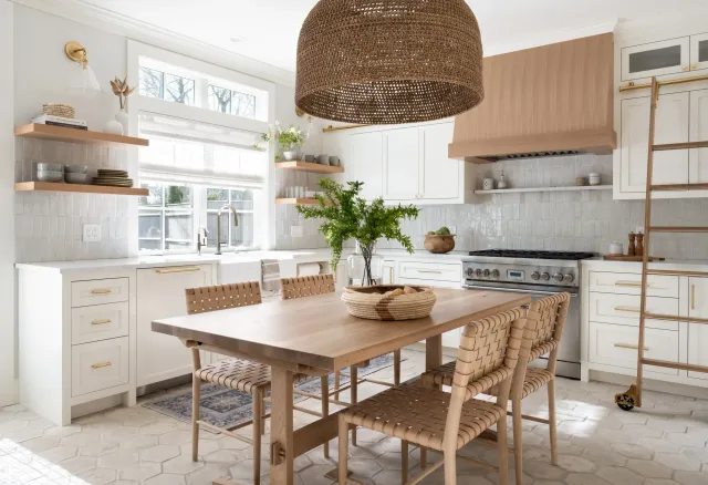 Bright kitchen with white cabinetry, wooden accents, and a wooden dining table with woven chairs. Large window, green plants, and open shelves with dishes and decor.