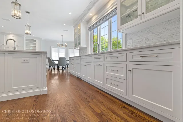 White kitchen with large windows, white cabinetry, and hardwood floors. A dining table with chairs is visible in the background, and the kitchen has modern lighting fixtures.