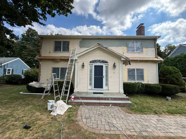 Two-story house under renovation with siding partially removed, ladders, and construction tools in front yard, cloudy sky in background.