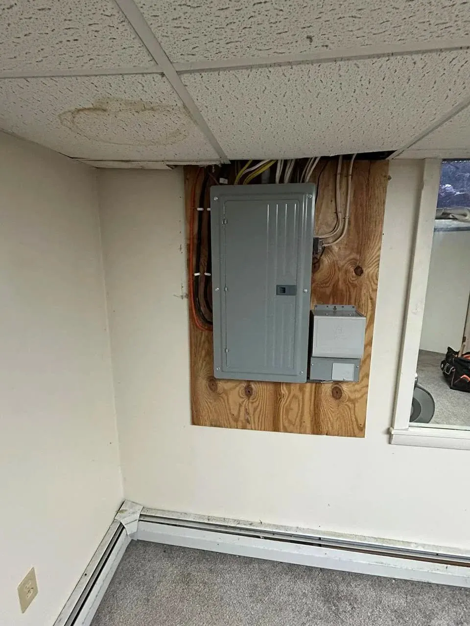 Electrical panel on wooden board in a room with drop ceiling tiles, showing water damage.