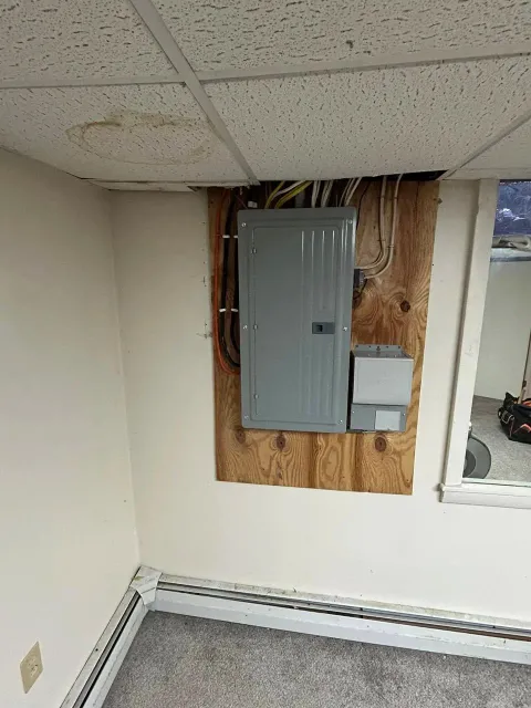 Electrical panel on wooden board in a room with drop ceiling tiles, showing water damage.
