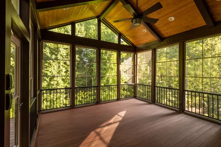 A screened-in porch with wood flooring, a high slanted wood ceiling, a ceiling fan, and views of surrounding trees.