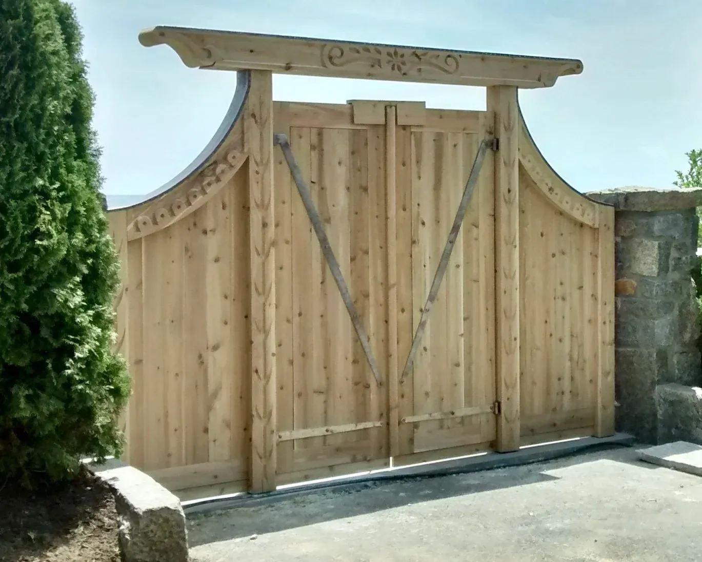 A wooden sliding gate with decorative carved details, mounted on metal tracks, flanked by a stone wall on the right and a bush on the left.