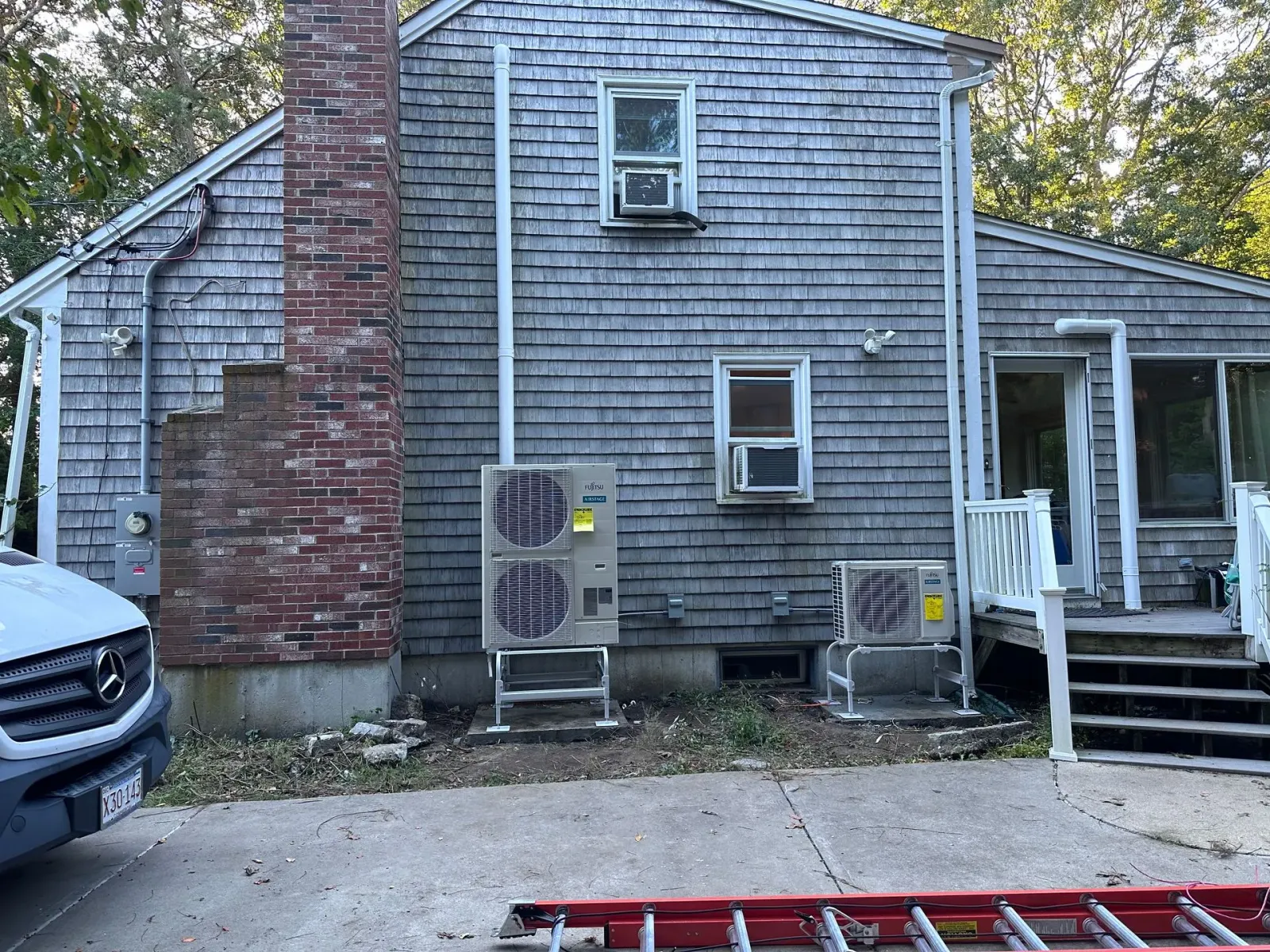 Back of a gray shingle house with brick chimney, two air conditioners, and a red car in the foreground