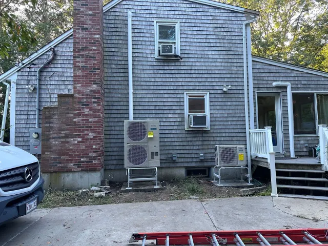 Back of a gray shingle house with brick chimney, two air conditioners, and a red car in the foreground