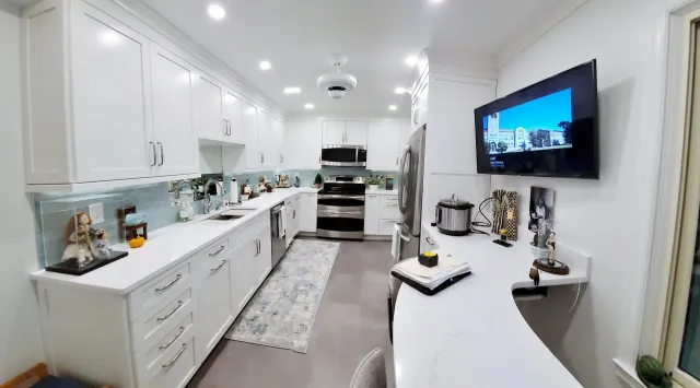 A kitchen with white cabinets and stainless steel appliances and a flat screen tv hanging on the wall.