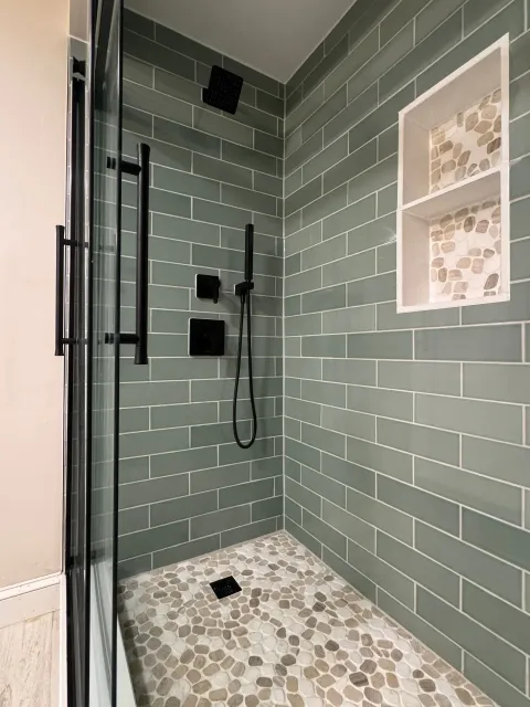 Walk-in shower with green subway tile walls, pebble floor, black fixtures, and built-in shelf niche