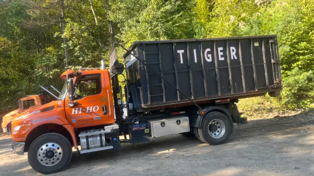 Orange trash collection truck with black dumpster labeled 'TIGER' parked on a dirt road surrounded by green trees.